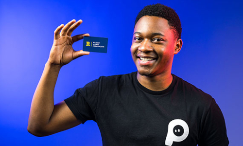 Tamuda Chimhanda holds a business card with the Rochester Alumni Ambassadors logo.